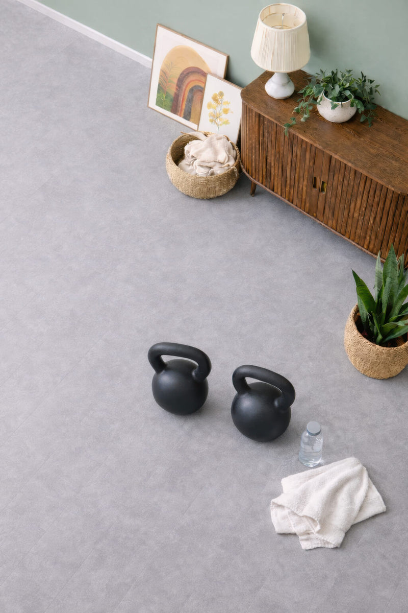 Two black kettlebells, bottled water, and a gym towel on Waterblock X Kasu grey stone-look floor with a wooden cabinet and decorative items in the background.